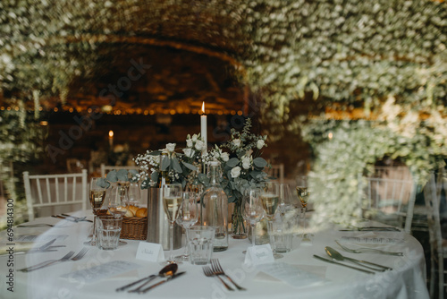 Elegant, romantic table setting for indoor wedding reception with fairy lights on wall behind. Luxury wedding table decoration, special event table set up with fresh flowers arragements. photo