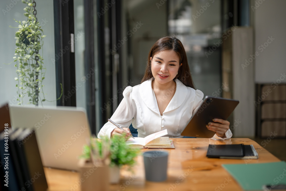 Asian business woman working using a digital tablet. Think, analyze data and plan work. Record in a notebook.