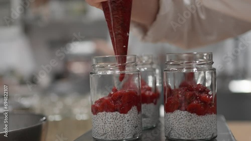 Employing a pastry bag, skillful hands carefully dispense strawberry jam into a jar, blending it with hydrated chia and fresh strawberries to craft a delightful dessert.