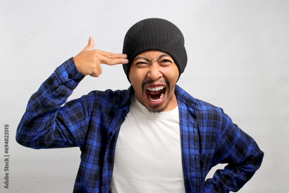 Distressed young Asian man, clad in a beanie hat and casual shirt, showing finger pistol gestures above his head, grappling with life challenges while standing against a white background. Distressed young Asian man, clad in a beanie hat and casual shirt, showing finger pistol gestures above his head, grappling with life challenges while standing against a white background.
