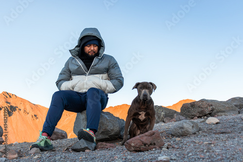 Man in warm clothes and in hood sitting on rock with pet dog in daylight photo