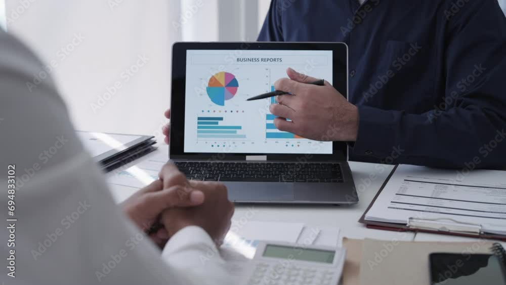 Asian businessman conversing with a team colleague, presenting and discussing a project via laptop. Engaged in purposeful work within a modern office setting. Professional collaboration.