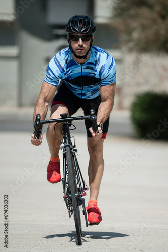 Front view of muscular young man cycling in a city looking at camera. Athletic cylcist training and riding a bike wearing helmet and sunglasses in the street. photo
