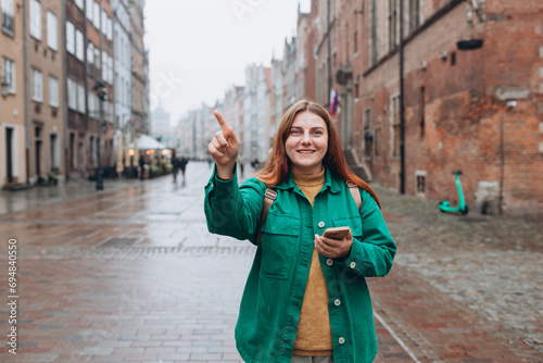Happy cheerful young woman with phone walking on city street checks her smartphone. Redhead girl with backpack pointing finger on city street. Traveling Europe in autumn. Urban lifestyle concept.