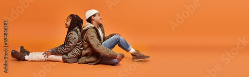 preteen african americna children sitting back to back on floor on orange backdrop, banner photo