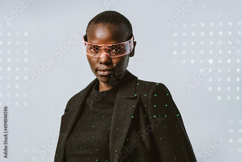Portrait of confident young bold African American woman in smart futuristic VR glasses and long black trench coat standing looking at camera against neon light illuminated background photo