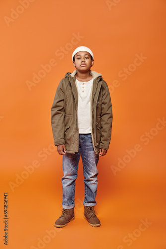 preadolescent african american boy in winter attire with beanie hat posing on orange backdrop
