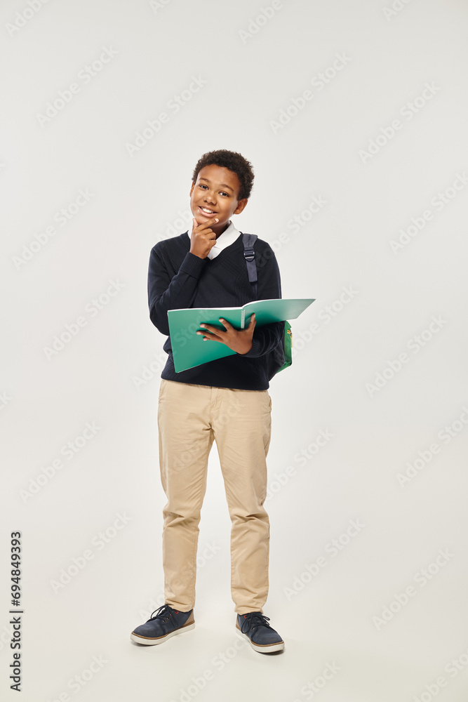 happy african american schoolboy in uniform holding notebook on grey background, hand near chin happy african american schoolboy in uniform holding notebook on grey background, hand near chin