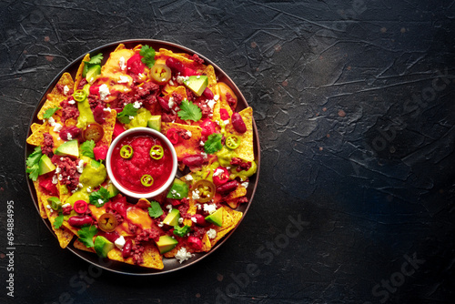 Loaded nachos. Mexican nacho chips with beef, overhead flat lay shot with guacamole sauce, cheese salsa, and chili peppers, on a black slate background, with a place for text
