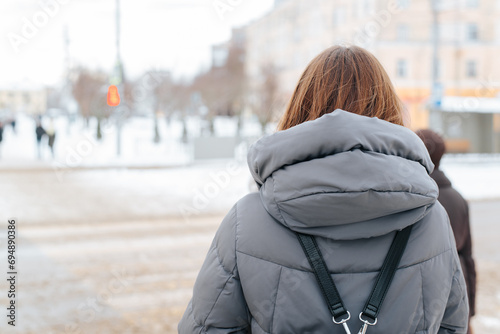 Back view of a female pedestrian in winter clothes standing by the road at a traffic light
