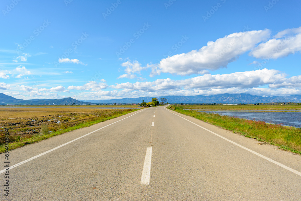 A straight road leading towards mountains under a bright blue sky with clouds, view of lonely road in the ebro delta, tarragona, catalonia, spain
