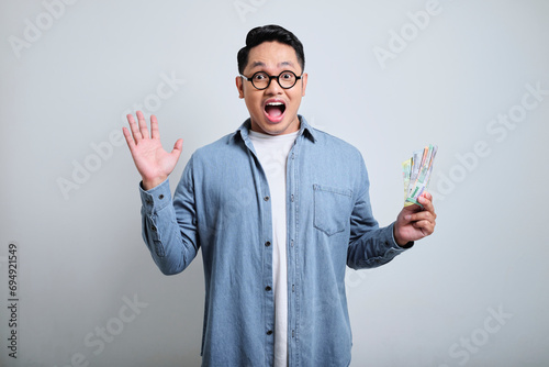 Portrait of handsome young asian man wearing denim and glasses in the smile face holding money and raising his right hand up, excited and happy, look at the camera.