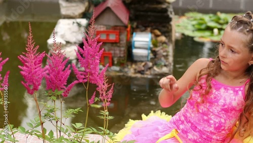 little girl sits next to pond and looks at flowers and bee photo