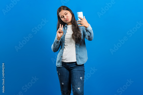 Image of an Indian women showing v-sign pose and sending air kiss while capturing self portrait photograph on blue background
