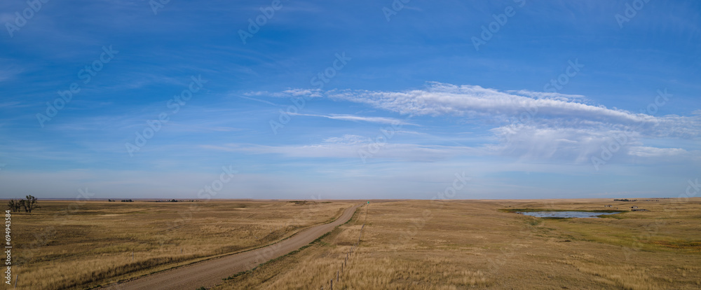 View of an extensive blue sky with scattered white clouds above a dry grassland prairie setting. A rural gravel road and a fence line cross the image and disappear into the distance.
