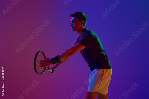 Competitive man in his 30s, tennis player in motion, serving ball with racket, practicing against gradient studio background on neon. Concept of professional sport, competition, game, math, hobby