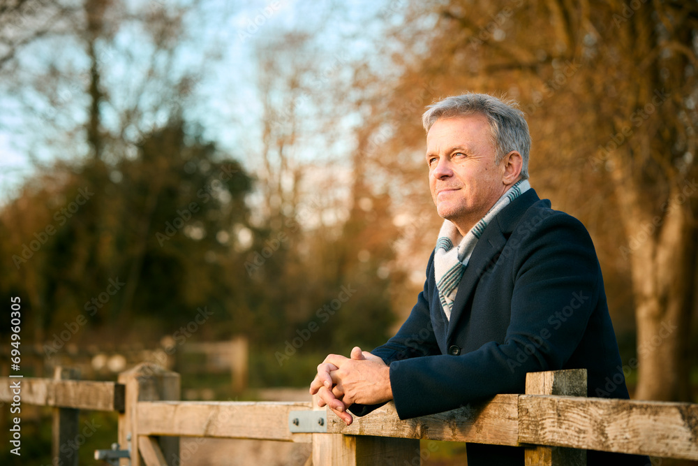Fototapeta premium Smiling Senior Man Leaning On Wooden Fence On Walk Through Autumn Park Or Countryside
