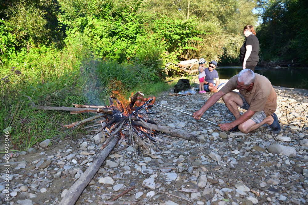 The family is camping by the river and roasting sausage over a natural fire