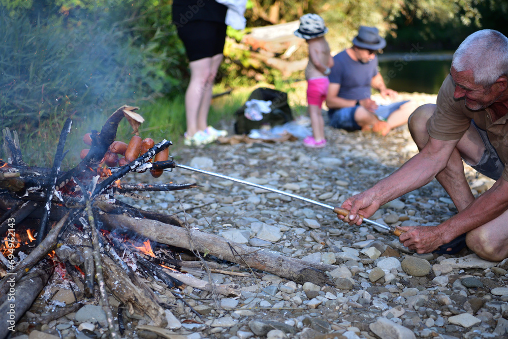 The family is camping by the river and roasting sausage over a natural fire