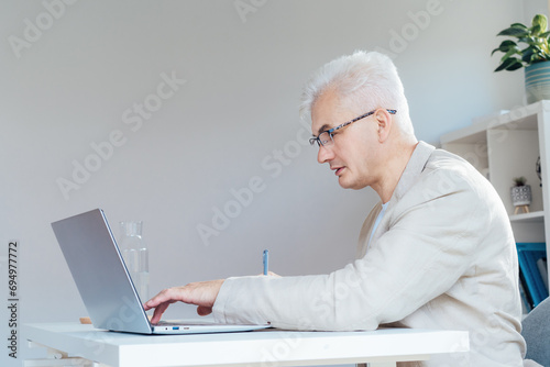 Concentrated grey hair middle-aged man making notes while working on laptop at work place in home office. Confident, experienced senior male professional. Small business entrepreneur manage business.