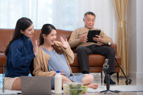 A pregnant asian woman sits on the floor with her younger sister. Live social media broadcast selling pregnant mothers' products and meals, There is a father in the background relaxing on a sofa.