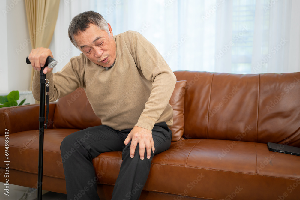 With knee joint pain, a senior guy sits on a sofa with a walking stick in his living room at home.