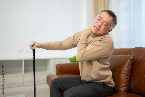 With shoulder pain, a senior guy sits on a sofa with a walking stick in his living room at home.