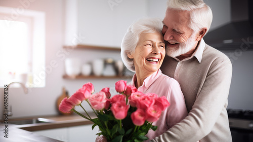 Elderly couple in a kitchen, with the man hugging the woman from behind, both smiling joyfully, as the woman holds a bouquet of roses. © MP Studio