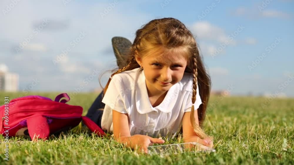 Little girl with pink backpack lies with tablet pc on green field Little girl with pink backpack lies with tablet pc on green field