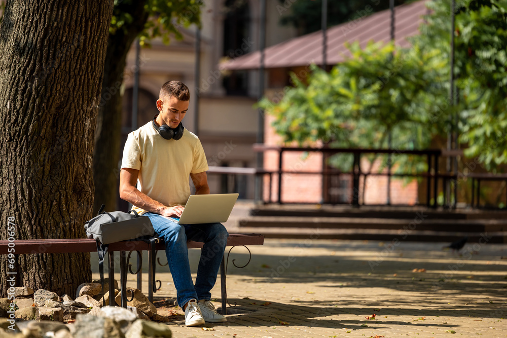 Obraz premium Short-haired young guy sitting under the tree and working on a laptop