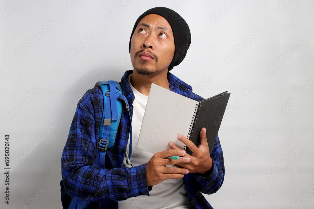 A pensive young Asian student, dressed in a beanie hat and casual shirt, carrying a backpack and holding a notebook, is engrossed in tackling a challenging assignment, deep in thought, exploring ideas A pensive young Asian student, dressed in a beanie hat and casual shirt, carrying a backpack and holding a notebook, is engrossed in tackling a challenging assignment, deep in thought, exploring ideas