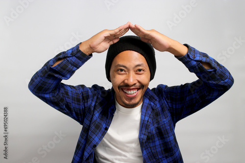 Happy Young Asian man with a beanie hat and casual clothes is making a hand gesture above his head, symbolizing a roof or home while standing against white background photo