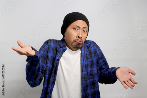 Confused young Asian man, dressed in a beanie hat and casual shirt is shrugging his shoulders in a gesture of cluelessness while standing against white background. photo