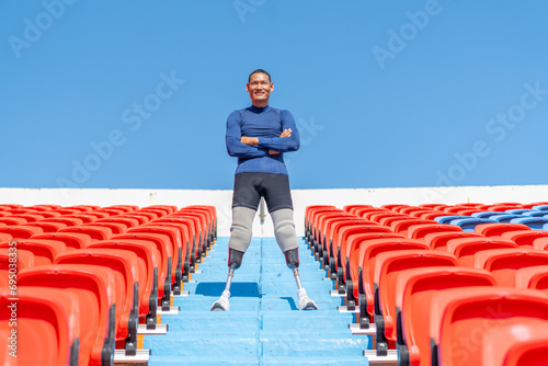 Sport man athlete prosthesis legs stand with arm crossed on stairs with happiness also look at camera on amphitheater in the stadium with day light. photo