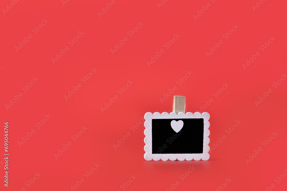 Small white board, placed on one side of image, held by wooden clothespin with white heart inside. Love. Red background. Horizontal.