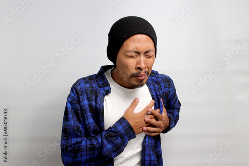 Fotografie Young Asian man in a beanie hat and casual shirt is clutching his chest, displaying symptoms consistent with a possible heart attack