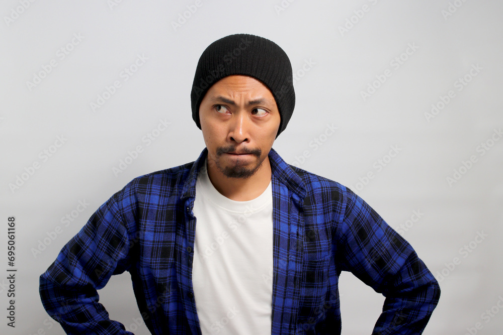Confused young Asian man is pursing or pinching his lips, displaying doubt and uncertainty while refraining from sharing important information. He appears hesitant, glancing sideways at copy space Confused young Asian man is pursing or pinching his lips, displaying doubt and uncertainty while refraining from sharing important information. He appears hesitant, glancing sideways at copy space