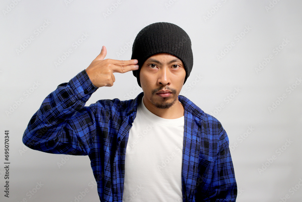 Distressed young Asian man, clad in a beanie hat and casual shirt, showing finger pistol gestures above his head, grappling with life challenges while standing against a white background. Distressed young Asian man, clad in a beanie hat and casual shirt, showing finger pistol gestures above his head, grappling with life challenges while standing against a white background.
