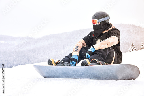 Preparing for a ride. Male snowboarder sitting with his equipment on the mountain and looking away