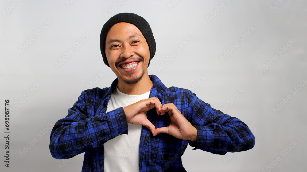 Attractive Asian man, dressed in a beanie hat and casual shirt, exudes happiness as he creates a romantic heart gesture with his hand, expressing tender feelings, standing against white background Attractive Asian man, dressed in a beanie hat and casual shirt, exudes happiness as he creates a romantic heart gesture with his hand, expressing tender feelings, standing against white background