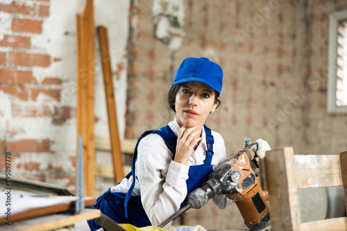 Focused female contractor wearing blue workwear standing with handheld demolition hammer inside building under construction photo