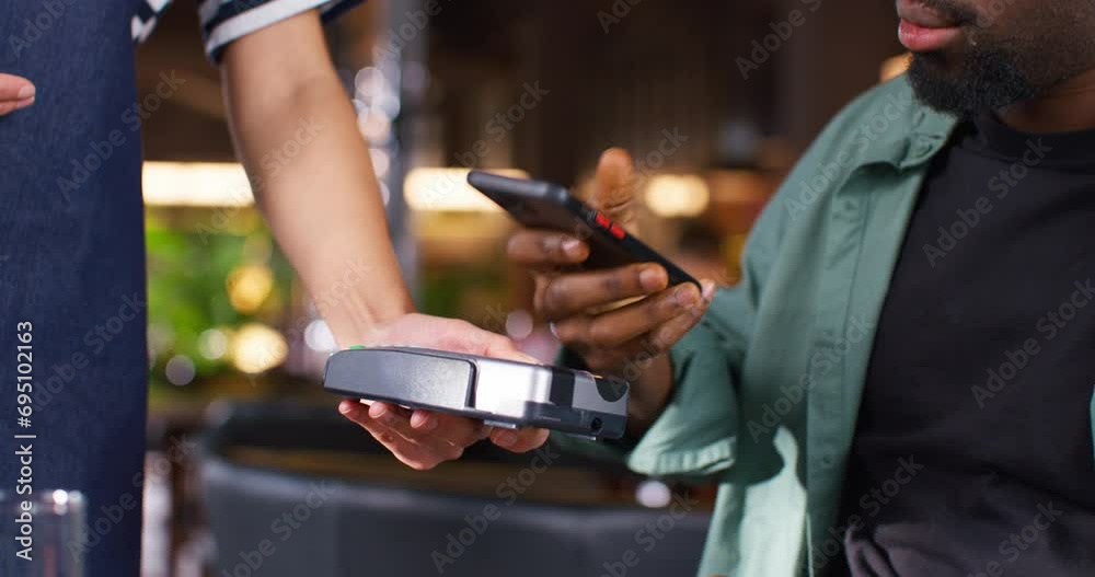 Camera view of waitress holding payment terminal while African American man paying with his telephone. Young guy using his phone for purchasing food or drink in cafe. Purchasing with technology device
