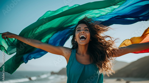 Fototapeta Naklejka Na Ścianę i Meble -  Joyful woman on the beach with arms outstretched, a colorful rainbow scarf flowing in the wind, with the ocean and blue sky in the background.