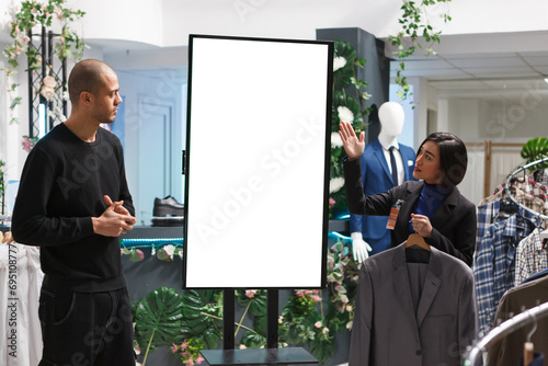 Boutique asian woman assistant pointing at empty marketing display mockup while helping arab man customer to choose clothes. Shopping mall store worker consulting client, showing brand advertising