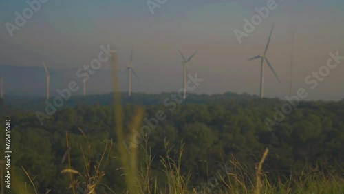 Wind turbines rise beyond tall grass under hazy blue sky, slender white blades turning across distant landscape, blending renewable energy with quiet rural scenery.