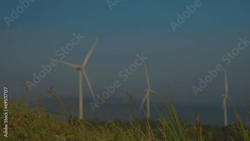 Wind turbines rise beyond tall grass under hazy blue sky, slender white blades turning across distant landscape, blending renewable energy with quiet rural scenery.
