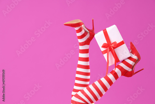 Female legs in Christmas stockings and high heeled shoes with gift box on color background