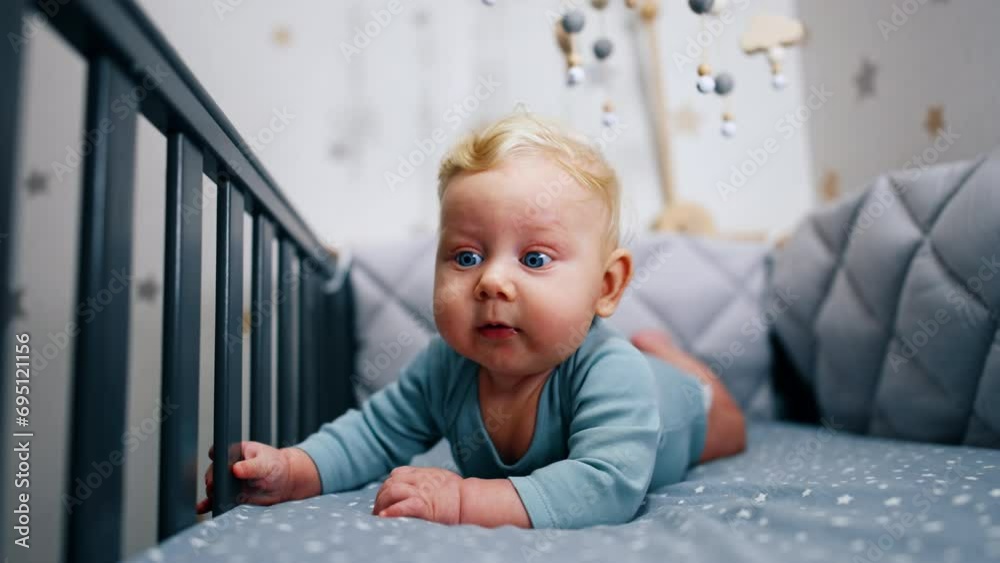 Blond Caucasian infant boy in blue bodysuit lies on the belly in the crib. Adorable blue-eyed boy looking up with surprise. Blond Caucasian infant boy in blue bodysuit lies on the belly in the crib. Adorable blue-eyed boy looking up with surprise.