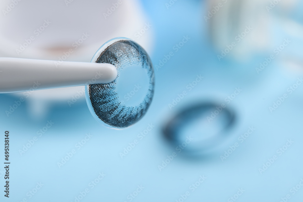 Tweezers with color contact lens on light blue background, closeup. Space for text - obrazy, fototapety, plakaty 