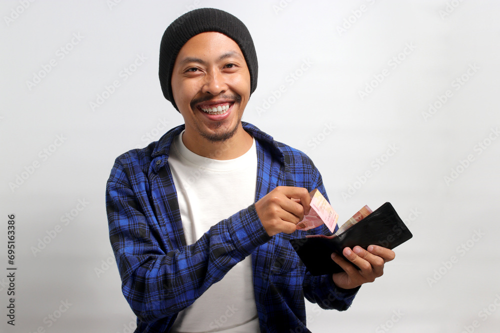 Excited Asian man, dressed in a beanie hat and casual shirt, smiles at the camera while proudly displaying a wallet filled with money, representing finance, investment, and money saving concepts Excited Asian man, dressed in a beanie hat and casual shirt, smiles at the camera while proudly displaying a wallet filled with money, representing finance, investment, and money saving concepts